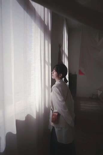 一个站在一间房间里的女人 透过薄纱窗帘向外看。