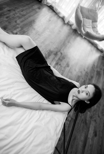 一个穿着黑色连衣裙的年轻女子躺在铺有白色床单的床上 窗外有窗户。