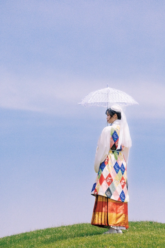 一位穿着连衣裙的女士手持白色雨伞站在绿草地上 背景是蓝天。