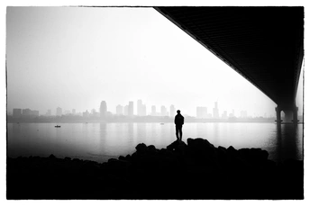 一张黑白照片 一个人站在桥下 面向水面。