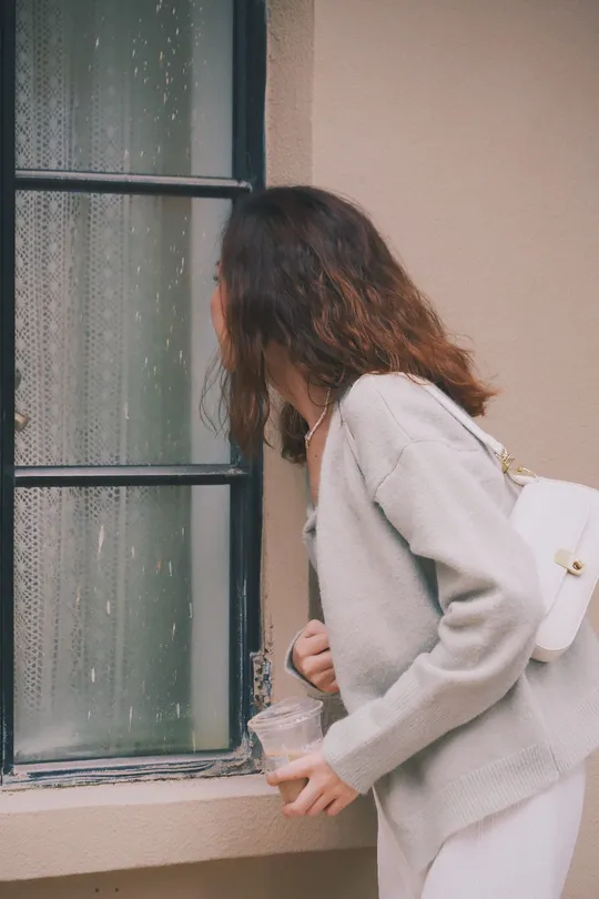 一个戴白色毛衣的女子正在窗户边拿着一杯东西