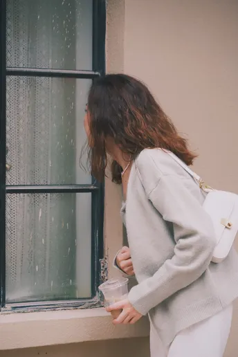 一个戴白色毛衣的女子正在窗户边拿着一杯东西