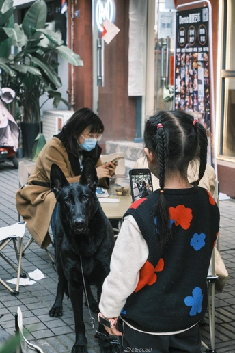 一个小女孩戴着口罩坐在桌子前 桌子前面有一只黑狗。