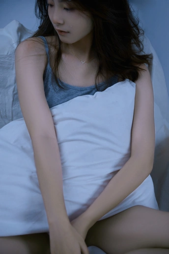 一个年轻女子坐在床边 床单和枕头都是白色的。