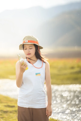 一个戴着帽子的小女孩正在从水瓶中喝水 她站在一条河边的田野上。