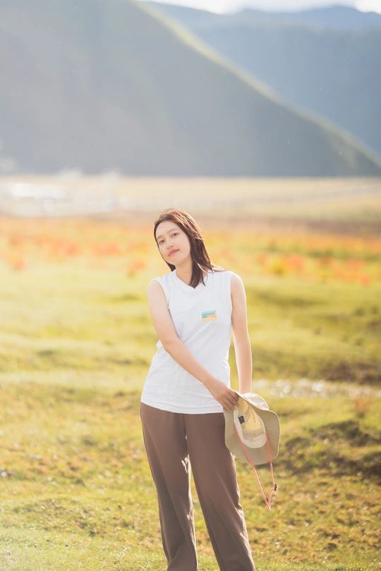 一位戴白色帽子穿着裤子的年轻女子站在有山脉背景的田野中