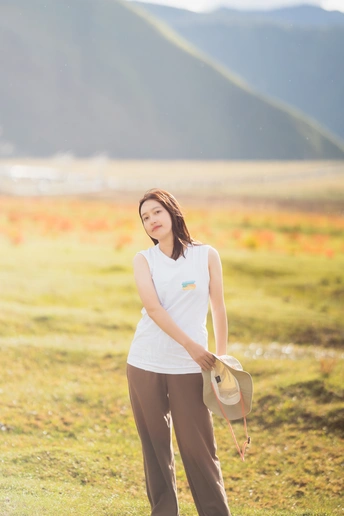 一位戴白色帽子穿着裤子的年轻女子站在有山脉背景的田野中