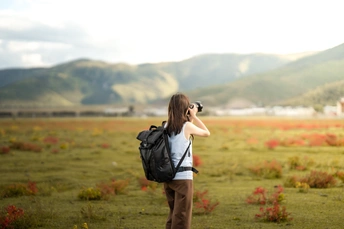 一位年轻女子背着书包站在一个有山脉背景的田野中 用她的相机拍照。