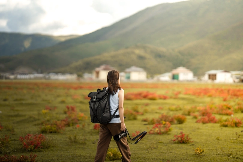 一位年轻女子背着书包和相机穿过一片花丛 背景是山脉。
