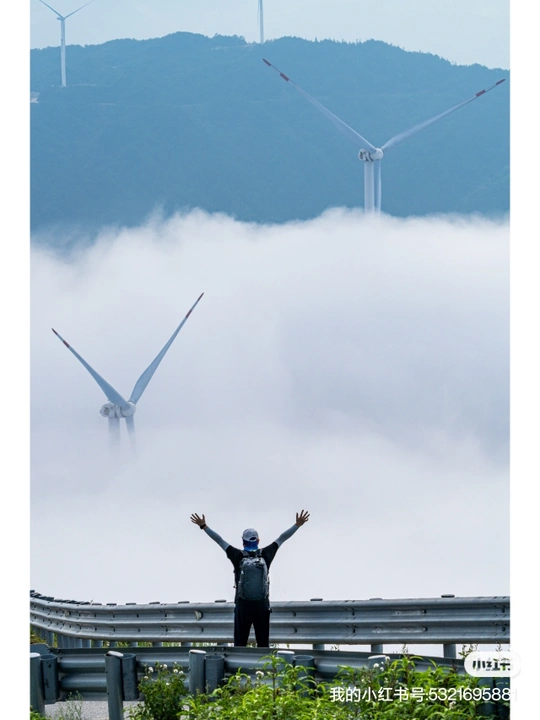 站在风力发电场前 伸展双臂的男子 背景是风车和被雾气笼罩的山峦。