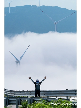 站在风力发电场前 伸展双臂的男子 背景是风车和被雾气笼罩的山峦。