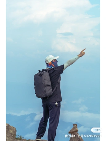 一个背着背包的男子站在山顶 双手举过头顶 手指向上指向天空。