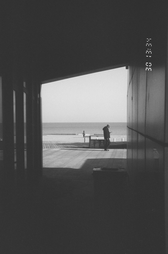 一张黑白的海滩照片 一个人站在长凳上 旁边有人在水边走。