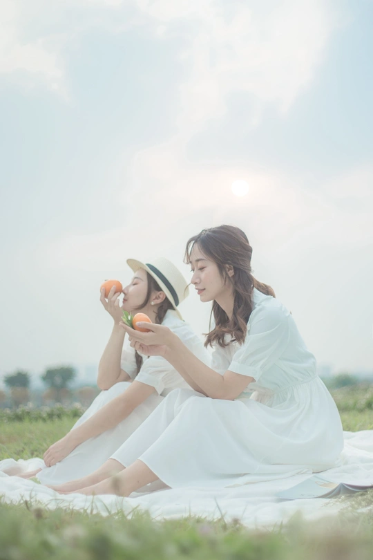 一个穿着白色连衣裙的年轻女子坐在草地上 她的母亲拿着一个苹果 一个年轻女孩坐在草地上。