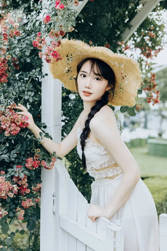 一位戴着草帽和白色连衣裙的美女站在围栏旁边 旁边有花。