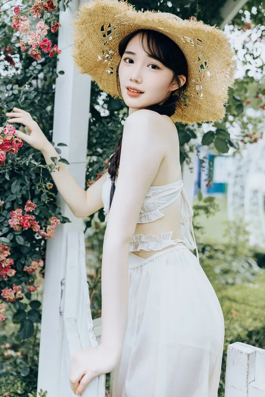 一位美丽的年轻女子穿着一件白色连衣裙和一顶草帽站在围栏旁边。