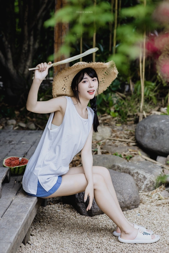 一位穿着白色连衣裙和草帽的年轻女子坐在木椅上花园里