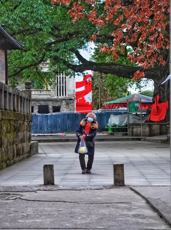 一位女士带着一个红色书包和一个小孩子走在树下的小街上