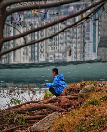 河边的岩石上坐着一个小男孩 背景是一座城市 前景是一棵树在水中的倒影。