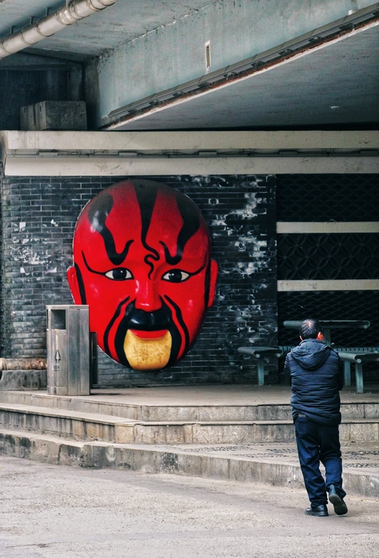 一个人站在一栋建筑前 头戴一个巨大的红色面具。