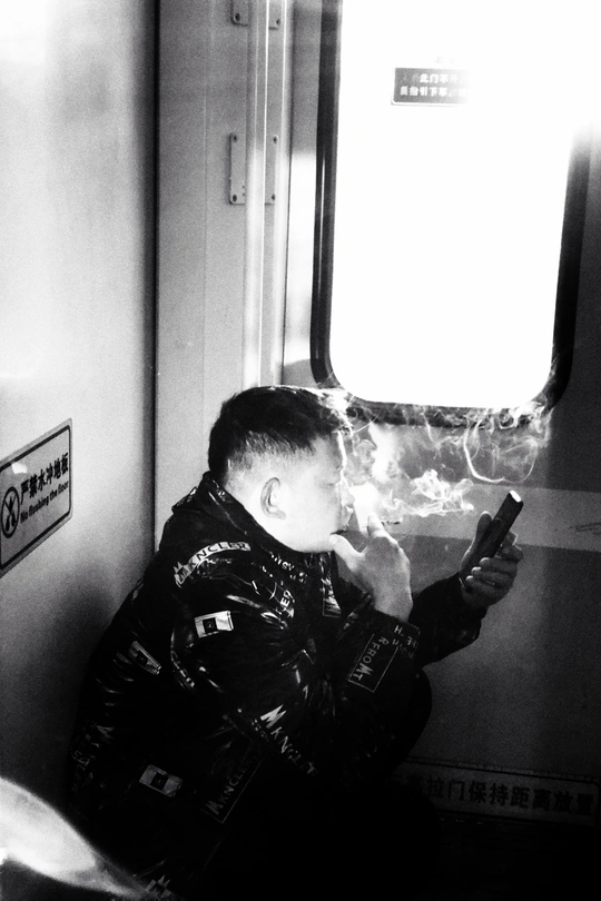 一张黑白照片 一个年轻男子坐在地铁上 正在看他的手机 抽烟。