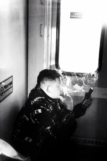 一张黑白照片 一个年轻男子坐在地铁上 正在看他的手机 抽烟。
