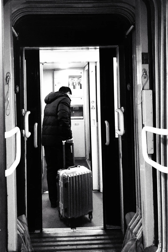 一个人站在一节火车车厢的走廊里 带着一个箱子 靠近门口。