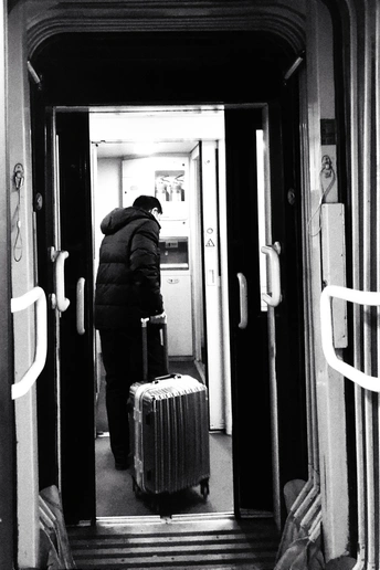 一个人站在一节火车车厢的走廊里 带着一个箱子 靠近门口。