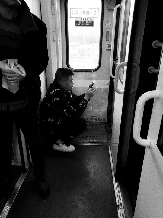 黑白照片显示一个人坐在地铁或地铁列车上 同时看着手机 而另一个人则坐在门上。