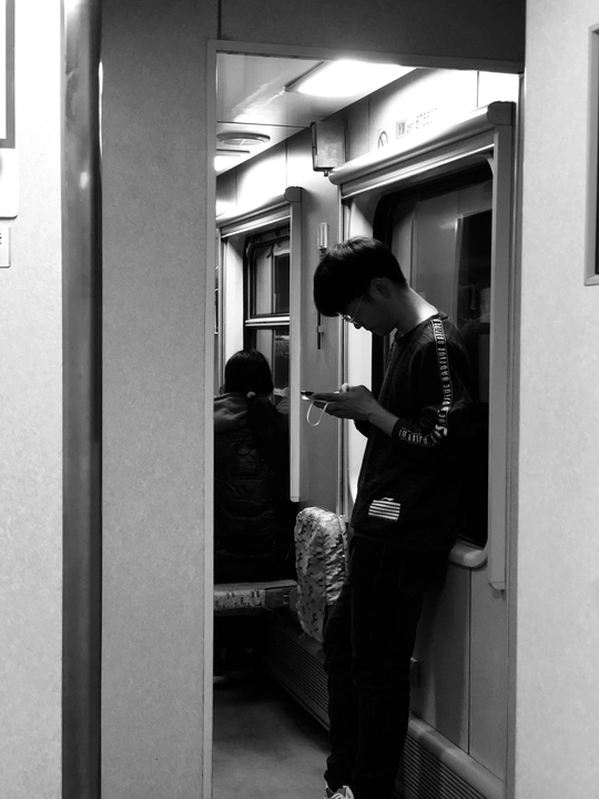一个年轻人在地铁列车上看着他的手机 而另一个人站在门口。