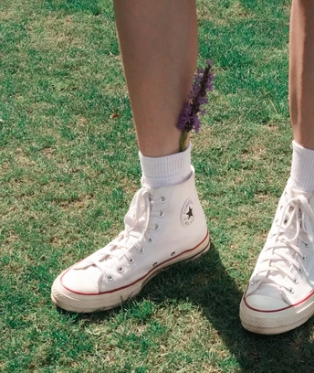 一个人站在草地上 穿着白色运动鞋和袜子 右脚上有一个紫色花。