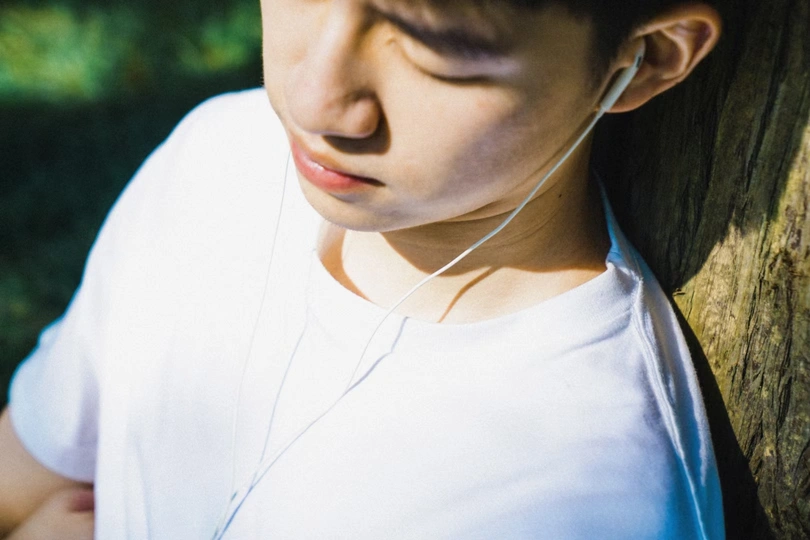 一个戴白色耳机、靠在树旁闭着眼睛的年轻男孩
