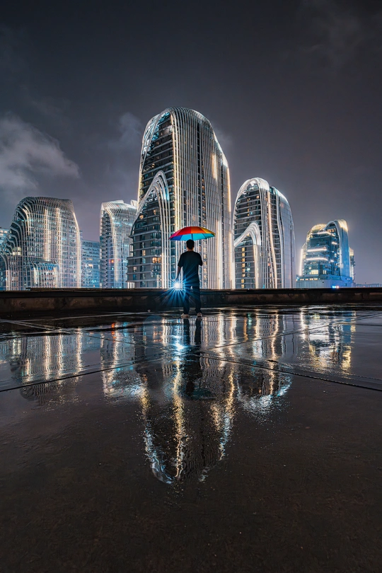 一位站在雨中 举着伞的女人 站在城市前面 背景是摩天大楼 水面上有倒影。