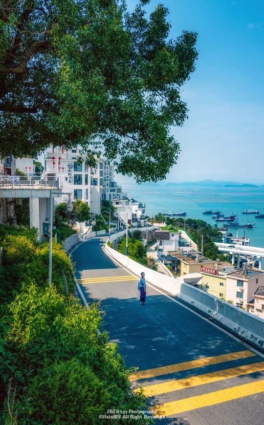 一个人沿着陡峭的山坡走下去 俯瞰着对面的公路和海港小镇。