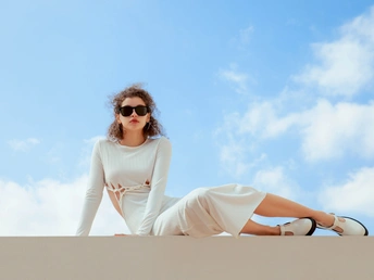 一个戴太阳镜和白色连衣裙的年轻女子坐在沙漠中的墙壁上 背景是蓝天。