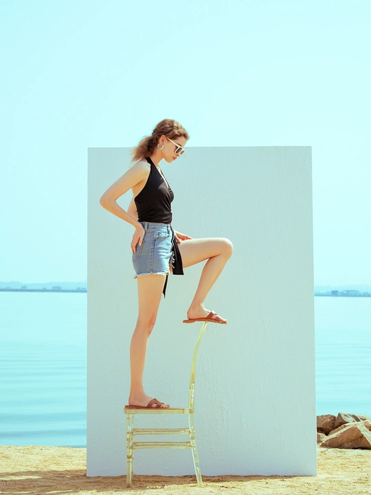 一位年轻女子站在一个沙滩上的小板凳上 倚靠在白色墙壁上 双腿交叉。