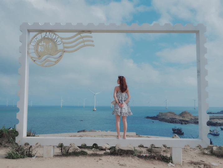 一张女人站在白色长凳上 眺望远方海面和远处风车的一张照片。