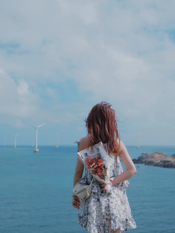 一位穿着连衣裙的年轻女子抱着花 从俯瞰海洋的悬崖上眺望着大海。