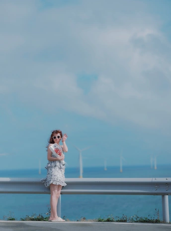 一个穿着连衣裙和太阳镜的年轻女孩站在桥上 俯瞰海洋和沙滩 空中飘着风筝。