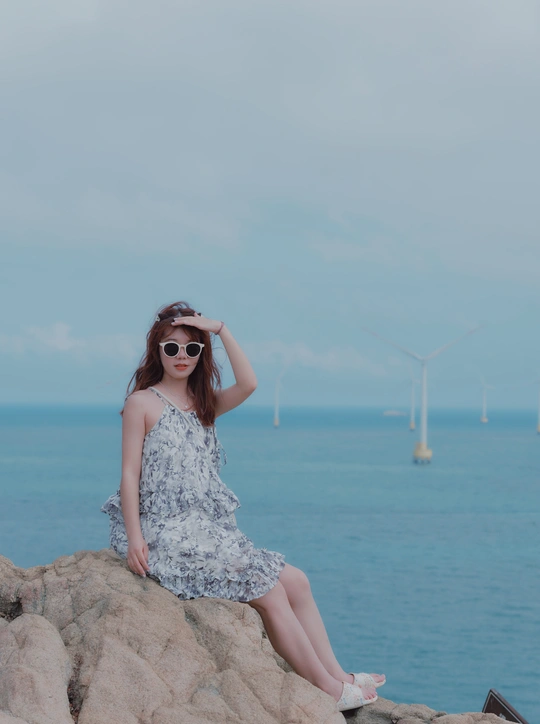 一个穿着连衣裙和太阳镜的年轻女子坐在俯瞰海洋和海面上的风车远处的岩石悬崖上。