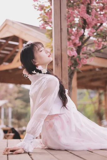 一位穿着白色连衣裙的年轻女子坐在地上 仰望天空 地板上有粉红色的樱花。