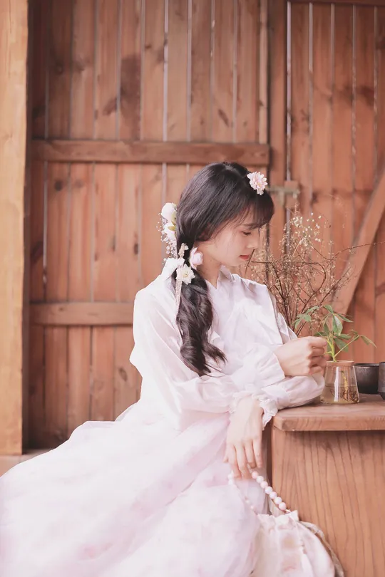 一个穿着传统服装的年轻女子坐在木结构建筑外面 手里拿着一束花。