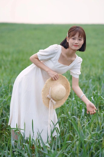 一位穿着白色连衣裙的年轻女子 戴着草帽站在一片绿草地上。