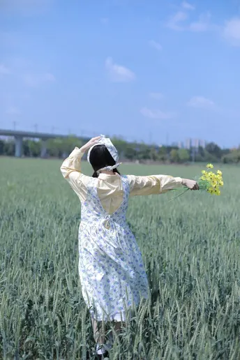一位穿着连衣裙的年轻女子站在绿麦田中 手中握着黄色花朵。