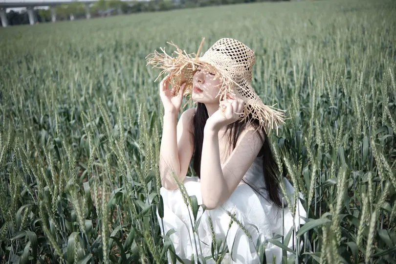 一个穿着连衣裙和草帽的年轻女子坐在一片碧绿的草地上 在一个美丽的麦田里。