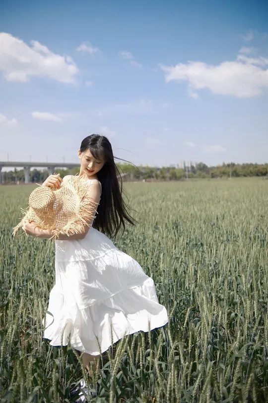 一位穿着白色连衣裙的年轻女子拿着帽子穿过一片麦田
