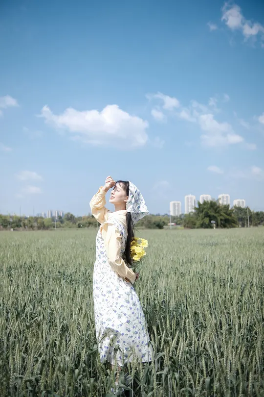 一位穿着连衣裙的年轻女子手持黄色花朵站在一片绿色麦田中