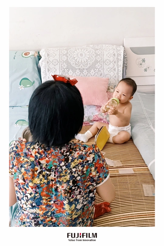 一个妇女坐在床边地板上 旁边有一个婴儿坐在地上 正在喝瓶中的东西。