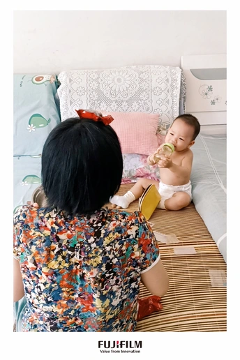 一个妇女坐在床边地板上 旁边有一个婴儿坐在地上 正在喝瓶中的东西。