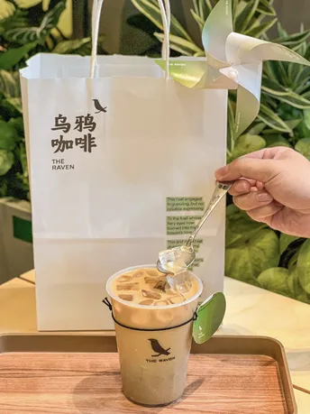 一个人在杯子旁边 手里拿着一根勺子 杯子里面装着咖啡 旁边还放着一个袋子。